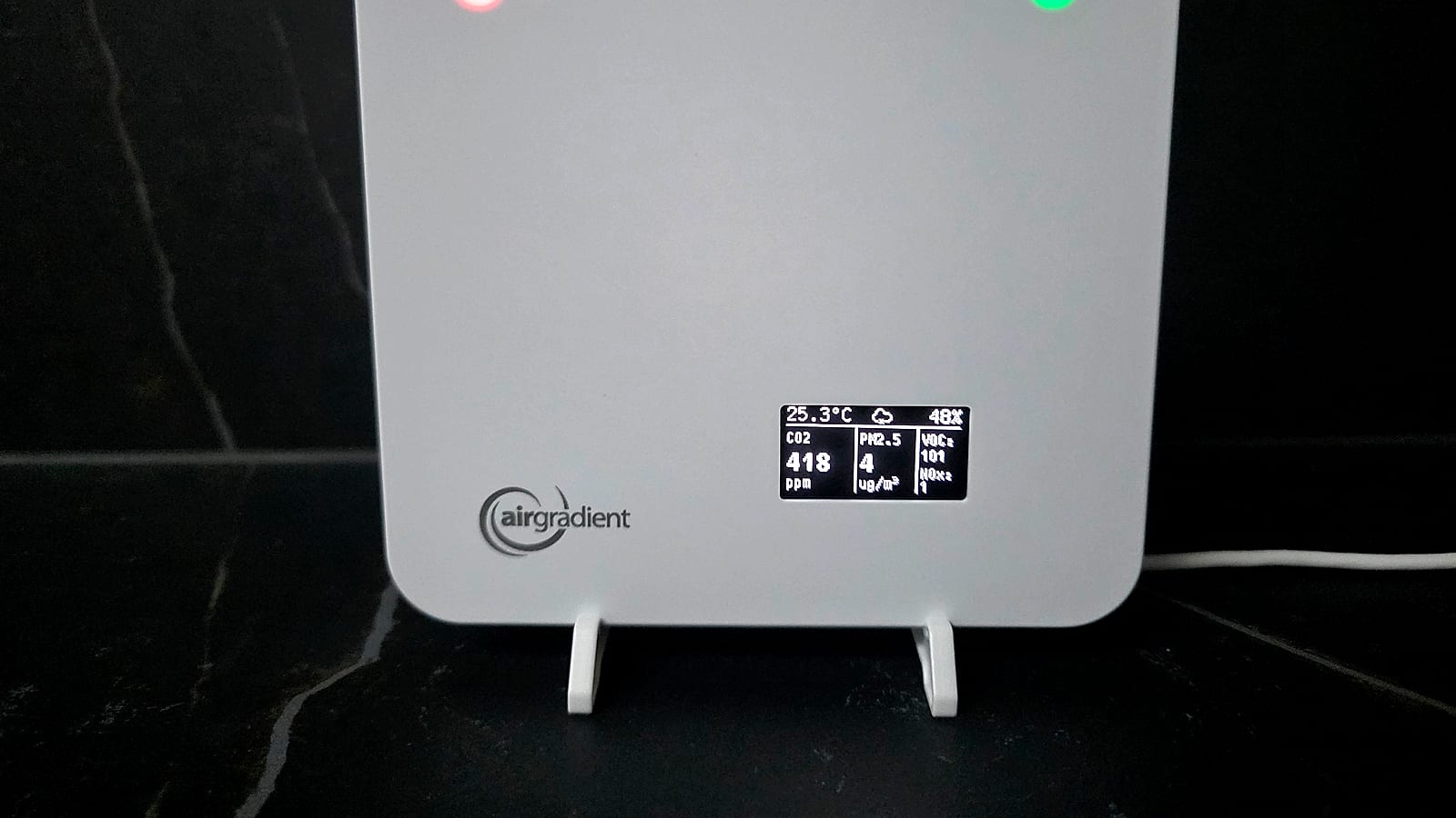 AirGradient ONE on a shelf with its OLED display and status LEDs lit, showing live CO2, PM2.5, VOC and NOx readings